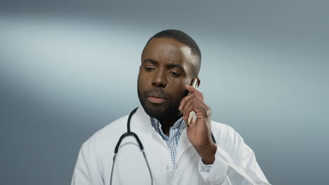 Close-up-of-the-African-American-young-male-doctor-or-intern-speaking-on-the-mobile-phone-on-the-gray-wall-background.